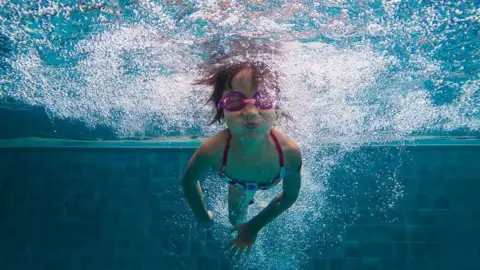A young girl swimming under water in a pool, she has pink goggles on and a bright swimsuit.