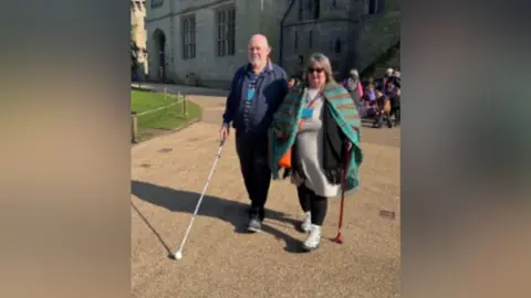 Warwickshire Vision Michael is on the left of the picture. He is wearing a blue top and black trousers and carrying a white cane with a ball at the end of his. His guide is next to him, with a green and red striped cape over a beige dress and black leggings.
