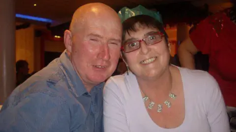 Family handout A picture of a couple. The woman is wearing red glasses, a necklace and a green paper crown. The man on the left is wearing blue shirt.