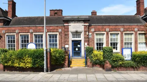 The building is a single level 1930's design with large rectangular windows and tall stone surrounded door which is painted blue.  There are yellow steps and blue railings up to the main door.