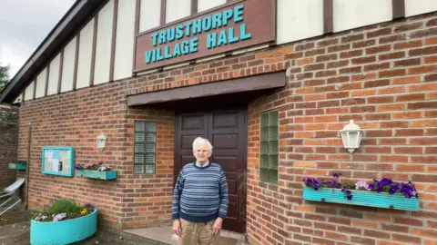 An elderly men with white hair, glasses, a stripy blue jumper and beige trousers is stood outside a village hall building. It reads Trusthorpe Village Hall in blue lettering with potted plants out the front.