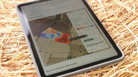 Janine Machin/BBC A tablet device sitting on a hay bale. Its screen shows a satellite image of a field with areas shaded blue and red in respect of where nitrogen is most and least prevalent.