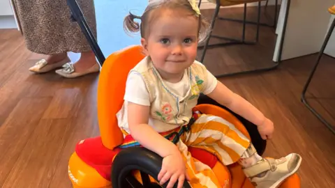 A young girl with blond hair, set into short pigtails, sits on a small, toy-like chair with wheels, which appears to have a handle at the back. It is set on a wooden floor and the feet of several stools can be seen behind her