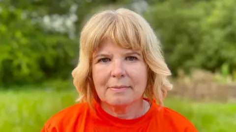 Nikki Fox/BBC Becci Butlin parada afuera en un espacio verde y mirando directamente a la cámara, tiene cabello rubio y viste una camiseta naranja.