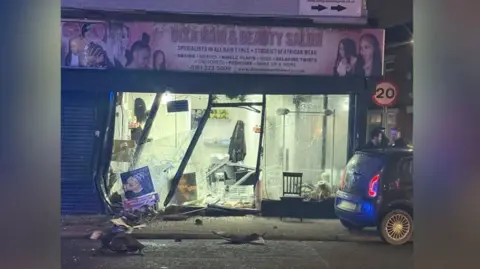 Smashed-up window at a beauty salon after a vehicle crashed into it. Glass and rubble lies in the road. It is dark but lights are still working in the damaged building.