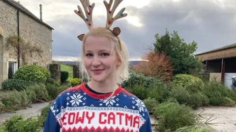 Anzhelika is wearing a festive blue sweater with cartoon cat faces and the text “MEOW CATMAS” in a Christmas-themed design. She has decorative reindeer antlers on her head. She is standing outdoors on a paved path surrounded by evergreen trees, with stone buildings and a cloudy sky in the background.