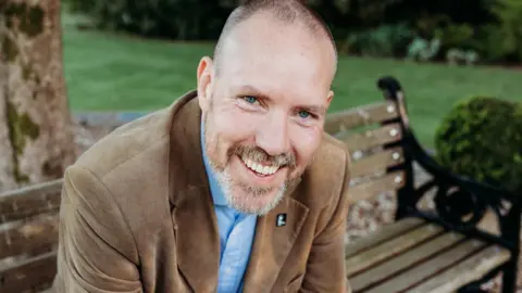 Graham, a man with a bright smile and grey stubble, who is wearing a brown jacket and blue shirt and sitting on a bench.