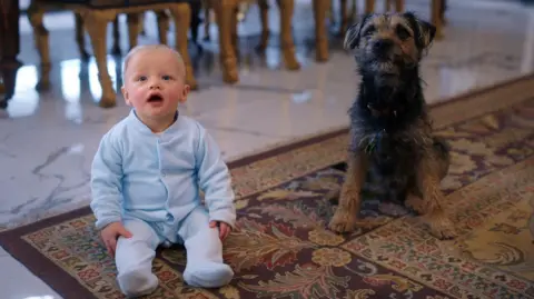 Netflix A close up of the baby and Artie sitting together on a traditional rug on a marble floor. The baby wears a pale blue sleepsuit and has blonde hair, and is looking up at someone taller. Artie sits on his hind legs with his two front legs out straight, lifting himself up a little and looking alert in the same direction as the baby. Artie's fur is a mix of black and reddish-brown.