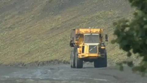 A yellow tipper truck at the opencast mine site. It is on flat, grey ground with a grassy slope behind it. 