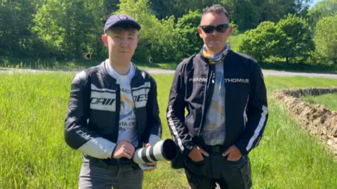 Both men are wearing biker gear with Ben holding a camera. Ben has a cap on and Jake is wearing sunglasses.