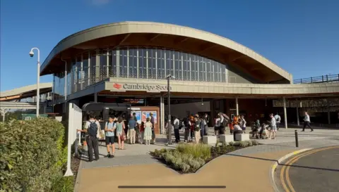 Greater Anglia A train station with an arched roof and people standing outside it.
It has a red sign which reads Cambridge South