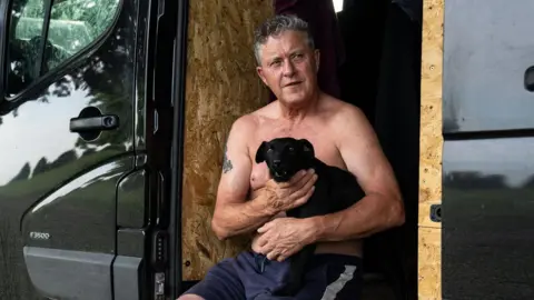 PA Media A man with no top on sits in the doorway of his van. He is holding a small black dog