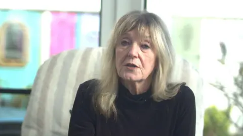 A woman wearing a black high neck long sleeve top sat on a cream chair. The background is blurred.