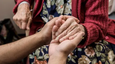 An older person is being comforted by somebody else who is holding their hands from outside the shot.