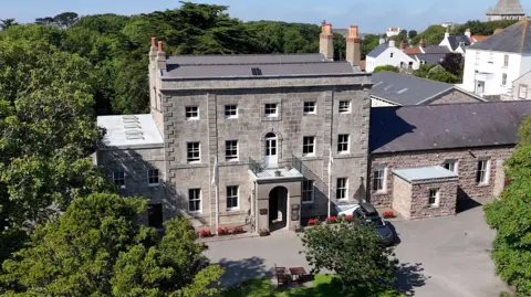 BBC A drone shot of the Alderney government building. It is a classic-looking grey brick building with small windows and a large porch entrance. It is surrounded by trees.