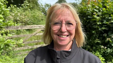 Andrea is outside on a sunny day. She is wearing a black, zipped jacket and has shoulder-length blonde hair and glasses. She is smiling at the camera. Behind her is a wooden farm-gate and hedges.