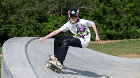 CGF Photography A boy skateboarding on a ramp on the side of a skatepark. He is wearing a black helmet, white T-shirt, black jeans and white trainers.