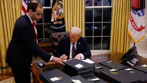 Getty Images Trump signs executive actions at the Resolute desk in the Oval Office. Piles of documents are on the desk, each with a Post-It note on saying what they are. An aide lifts a document as Trump uses a thick black pen to sign his name on another.