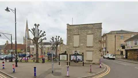 Centre of Higham Ferrers, showing a two-storey stone building in the centre, two bare trees to the right, a church steeple in the background, and two notice boards in front of the stone building. There are further stone buildings in the background and a road sweeping round to the right.
