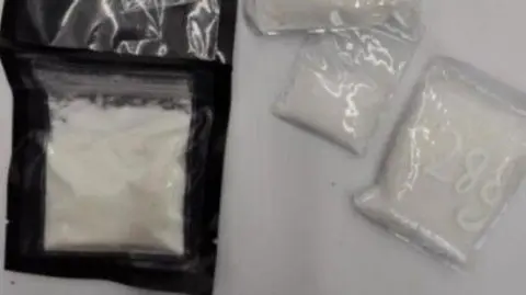 Sachets of the white powdered drugs on a white table. One of the sachets says 28 grams.