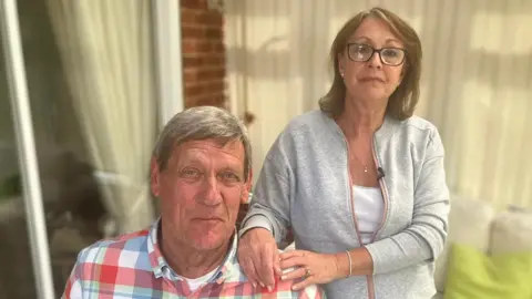 BBC Fiona has her hands on Nic's shoulder. They are both elderly, Nic has short grey hair and Fiona has long brown hair. He is wearing a checkered shirt and she is wearing a grey cardigan. 