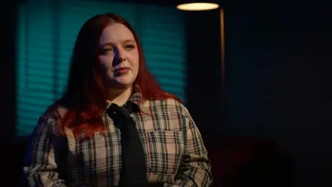 Amia Rohleder‑Cook sits during a filmed interview, wearing a checked shirt and tie, with blue lighting in the background.