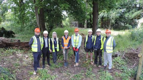 A group of people wearing hi-vis yellow and orange vests and standing on a patch of soil with shovels in the ground. They are wearing hard hats and smiling