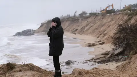 Joe Giddens/PA A woman in a black raincoat stands with her hood up and her phone to her ear with the sea in the background. Also in the background is the erosion-affected cliff with a digger on top demolishing one of the properties near the edge.