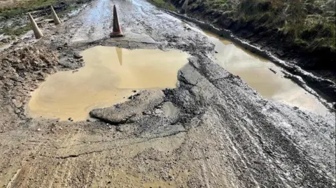 A muddy road which has two large potholes on it filled with muddy water and a grass verge on either side. There are also traffic cones in the road.