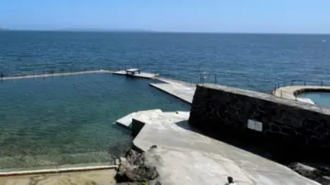 BBC The bathing pool at La Vallette which is sheltered from the open ocean with a stone wall. There is a low diving platform at the back of the pool. The sea is calm and blue. 