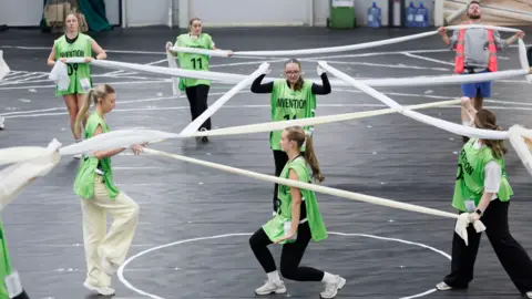 Glasgow 2026 A group of dancers wearing light green bibs, holding fabrics as they practice a dance routine 