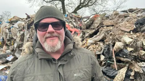 Jeremy Roney, a man wearing a green jacket with fur-lined hood and  a green woollen hat, standing next to a large pile of dumped waste material.