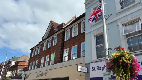 A lamp post on a suburban high street with a hanging flower basket and a Union flag. Shop fronts are in the background.