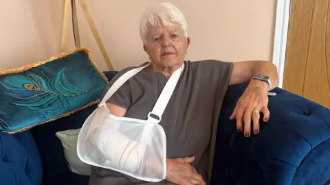 Ally Whyatt is sitting on a blue chair in her living room. She is wearing a brown top and trousers. On her right arm is a white sling supporting it, through the clear sling her bandaged injured arm is visible. Ally has white hair and is wearing pink lipstick.