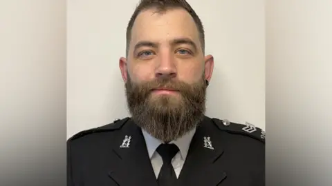 Warwickshire Police head and shoulders shot of Sgt Charlie Thompson, in his uniform. He has dark brown hair and similar coloured beard