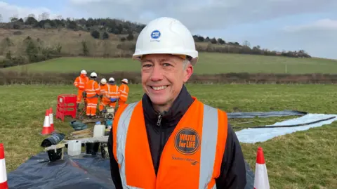 George Carden/BBC Joff Edervane wearing an orange Southern Water high vis vest and white hard hat smiling at the camera. Joff is standing in a field where the work is happening and behind him are five workers in the same clothes as him and there are tarpaulin sheets on the floor. The field is in the crest of a valley between two hills, one of which can be seen in the background