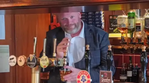 Stephen Cotterill A man with a greying beard, wearing a white shirt and a checked navy blazer, pulls a pint behind a wooden pub bar. In the background, shelves of spirits can be seen. 