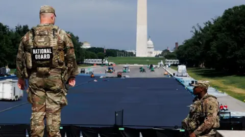 Two National Guards men in camouflage uniforms watch workers and trucks in the drained Lincoln Memorial reflecting pool, which is partially painted blue. The Washington Monument and the Capitol building are in the background