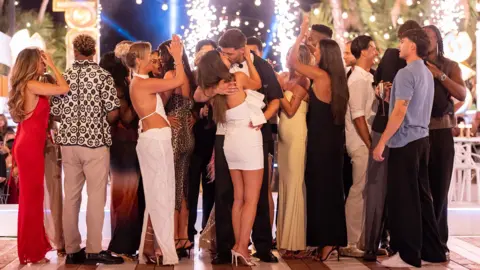 ITV Samie Elishi and Ciaran Davies share a kiss after they're announced as the winners of Love Island. It's a full length photo of all the couples stood on wooden decking on the Love Island set. Samie is wearing a white dress and white heels and Ciaran is wearing dark trousers and a dark shirt over a white t-shirt. They're both in the middle of the photo sharing a kiss while all the other islanders mingle and talk around them.