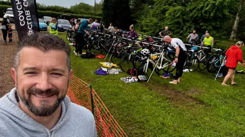 PA Media A man with short black and grey hair wearing a grey hoodie takes a selfie with people stood behind him in exercise clothing and racks of bikes. 