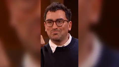 Dan Levy has short dark hair and is wearing glasses and a blue jumper.