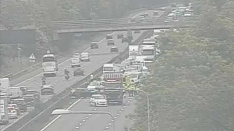 National Highways Cars are stopped on the road. Emergency service staff in high-vis jackets are standing on the road. There is a large queue of traffic behind the stopped vehicles.