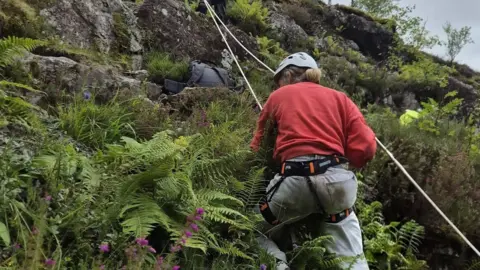 A woman in red jumper and grey trousers on ropes down the side of a hill in Glencoe.