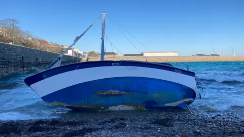 BBC A boat washed up along a rocky bay. The yacht is blue with a white stripe along it. There is a whole on the bottom of the vessel that looks like a rip and the sail has been completely ripped off. 