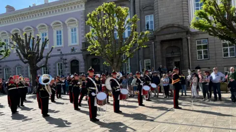 A marching band standing in the square outside the government building performing to a crowd standing in a long line on a sunny day.