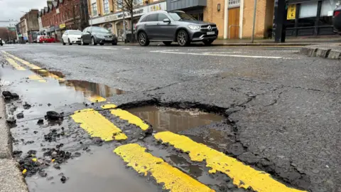Potholes filled with water on a road with double yellow lines. Across the street are a number of cars parked alongside shops. 