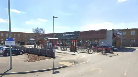 A Streetview image showing the McDonald's, which is situated in the corner of a large supermarket car park, surrounded on two sides by two storey buildings