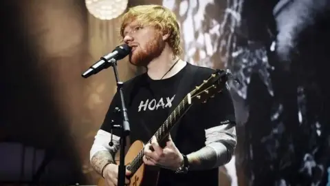 Getty Images Ed Sheeran plays his guitar and sings into a microphone on a stage. He has ginger hair and a beard and is wearing a black t-shirt which says 'hoax'. 