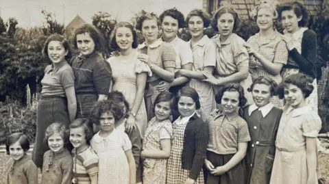 Jason Camis A black and white photograph of 19 girls ranging from about five years old to teenager years. They are standing in two rows, slightly side on, some with their hands on the shoulder of the girl in front. They're wearing an assortment of dresses, cardigans and short sleeved shirts in style during the 1940s. Most are smiling, but some look unsure.