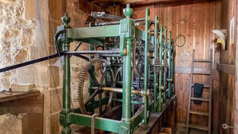 Anthony Davis A mechanism involving metal cogs and wheels which operates the clock in the tower of a church. There is a wooden ladder in the background propped up against a wooden wall. There is light coming in from the left from a window.